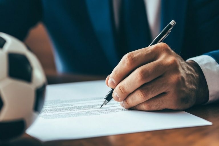 A man signing a piece of paper, with a mini football on the table in front of him.